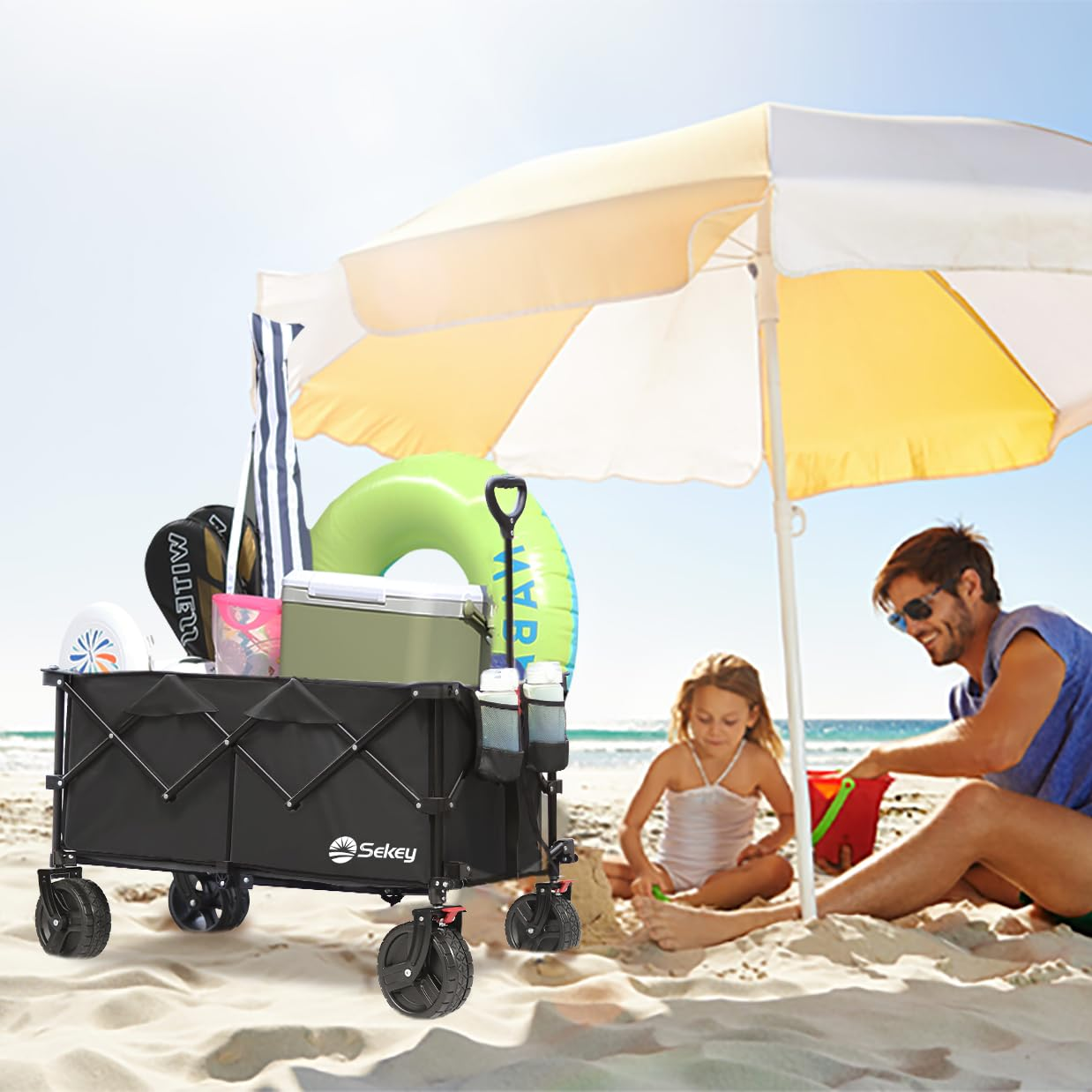 Un chariot pliant noir rempli d'équipements de plage est sur le sable près d'un grand parasol, d'un homme et d'un enfant.
