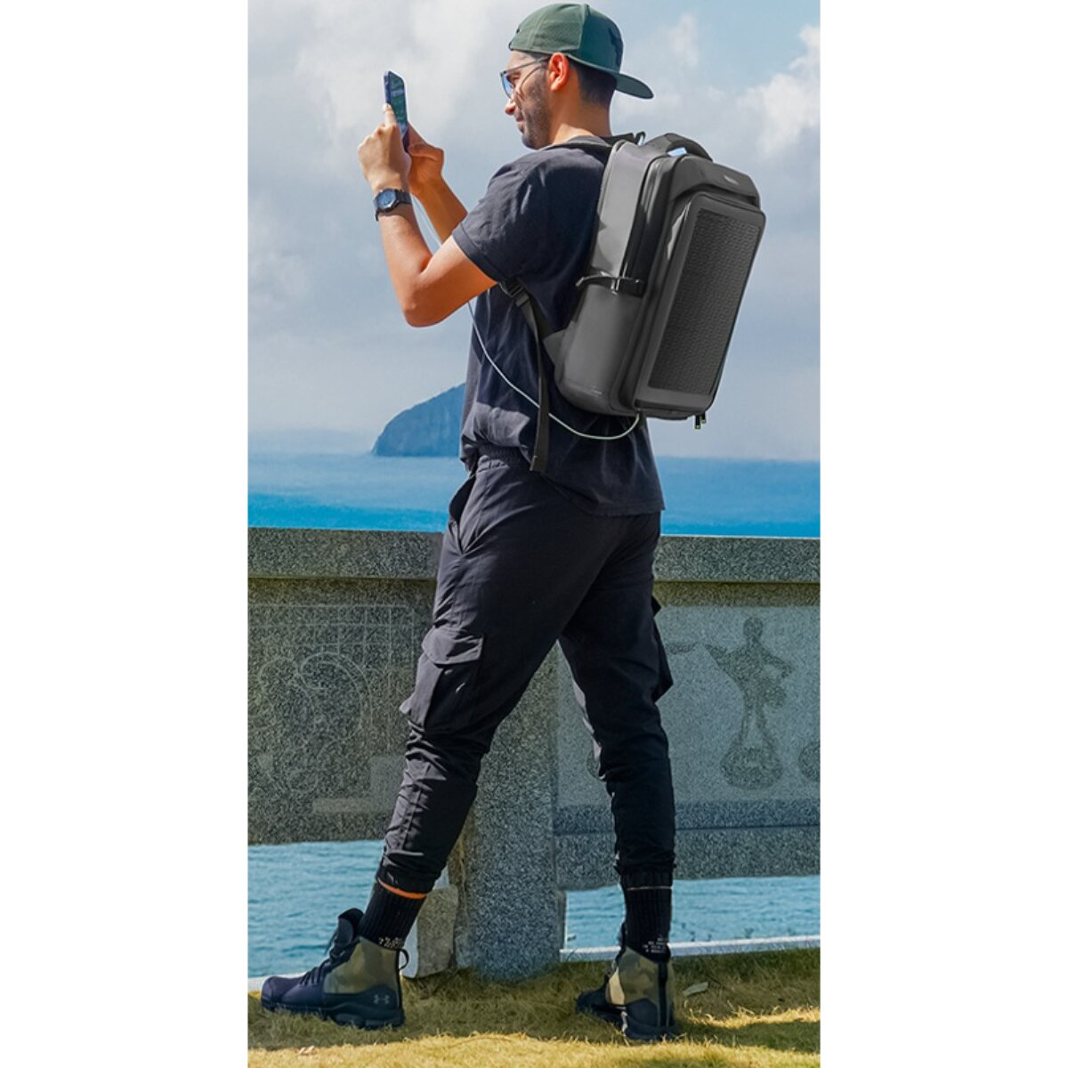 Ein Mann mit schwarzer Kleidung und Rucksack steht an einer Klippe und fotografiert das Meer mit seinem Smartphone.