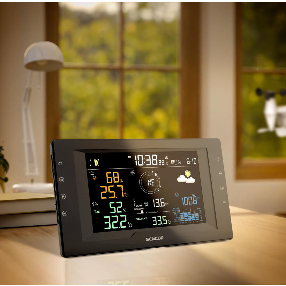 Eine schwarze Sencor-Wetterstation auf einem Tisch zeigt Uhrzeit, Temperatur, Luftfeuchtigkeit und Wettervorhersage an.