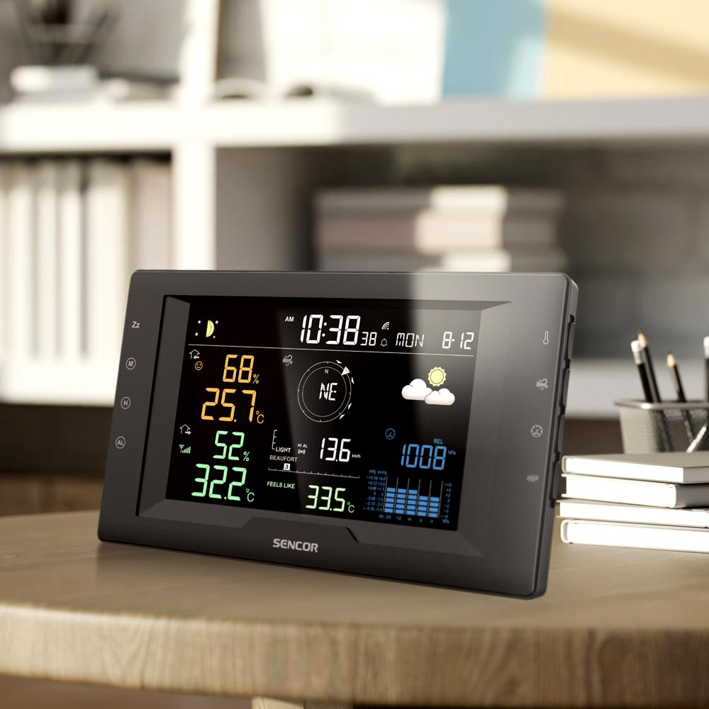 Digitale Wetterstation mit Farbdisplay auf einem Holztisch, im Hintergrund Regale.