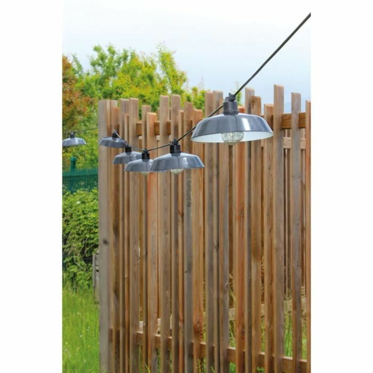 Eine Lichterkette mit grauen Industrie-Hängelampen hängt vor einem rustikalen Holzzaun im Garten.