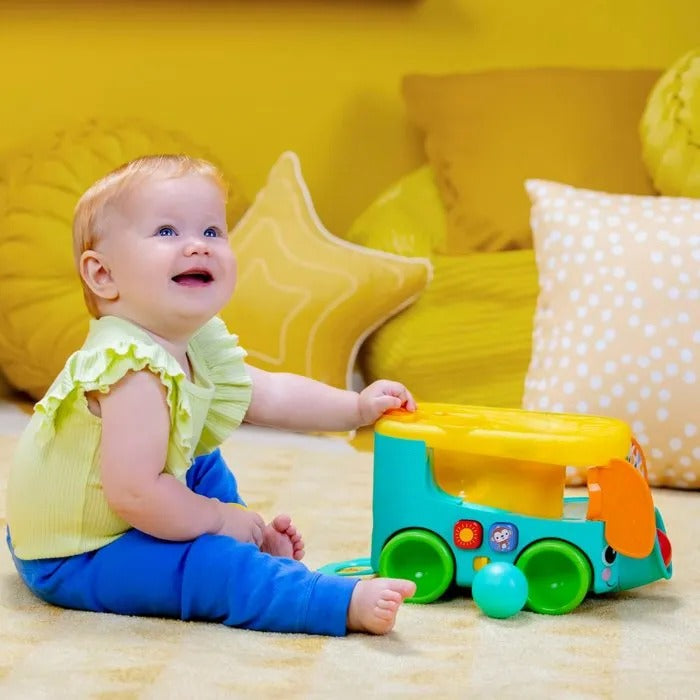 Ein lächelndes Baby sitzt auf dem Boden vor gelben Kissen und spielt mit einem bunten Spielzeugauto.