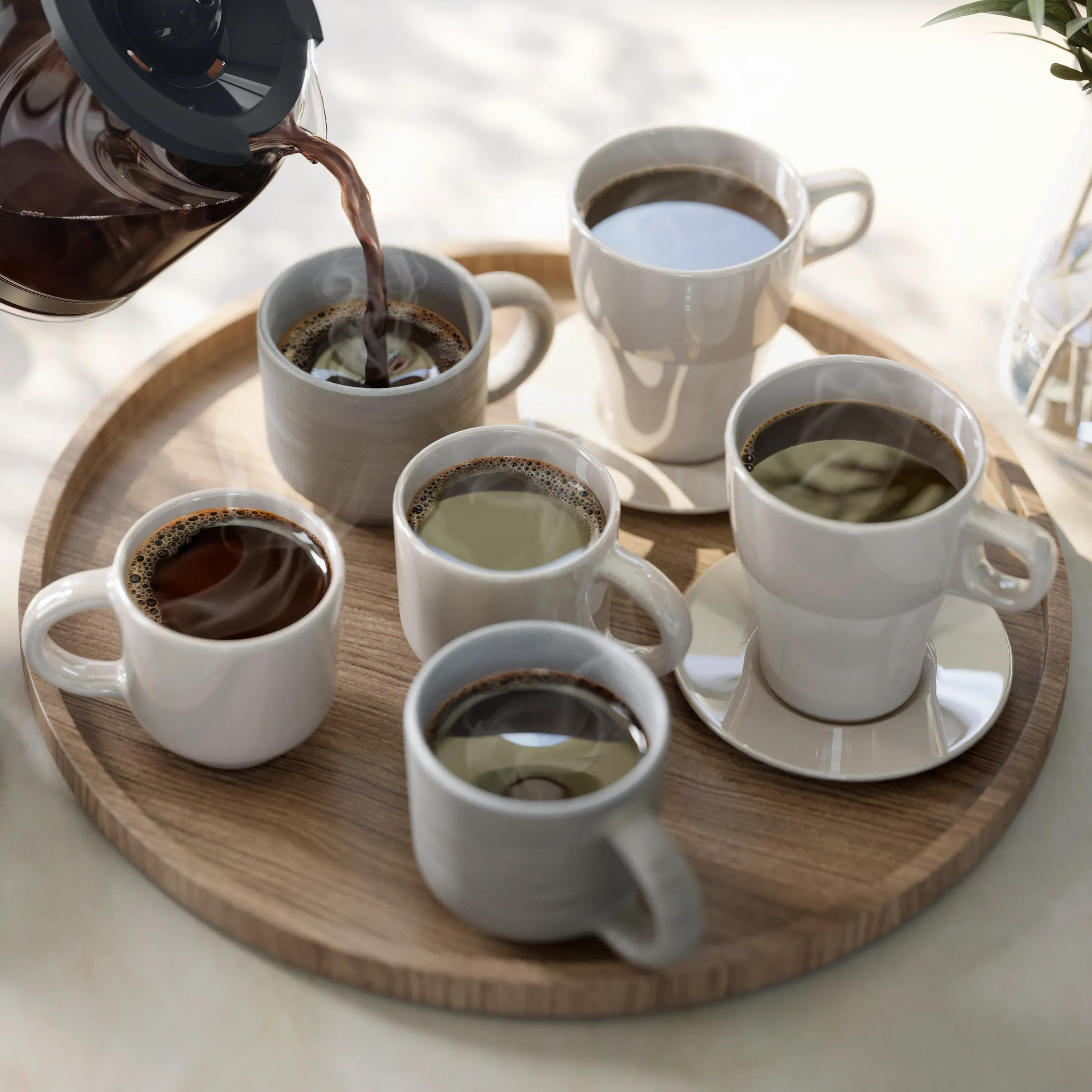 Coffee is poured from a pot into a cup on a wooden tray holding several other coffee cups.