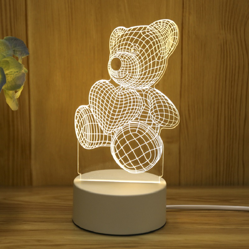 Eine beleuchtete 3D-Drahtgitter-Teddybären-Nachtlampe auf einem weißen runden Sockel vor einem Holzhintergrund.