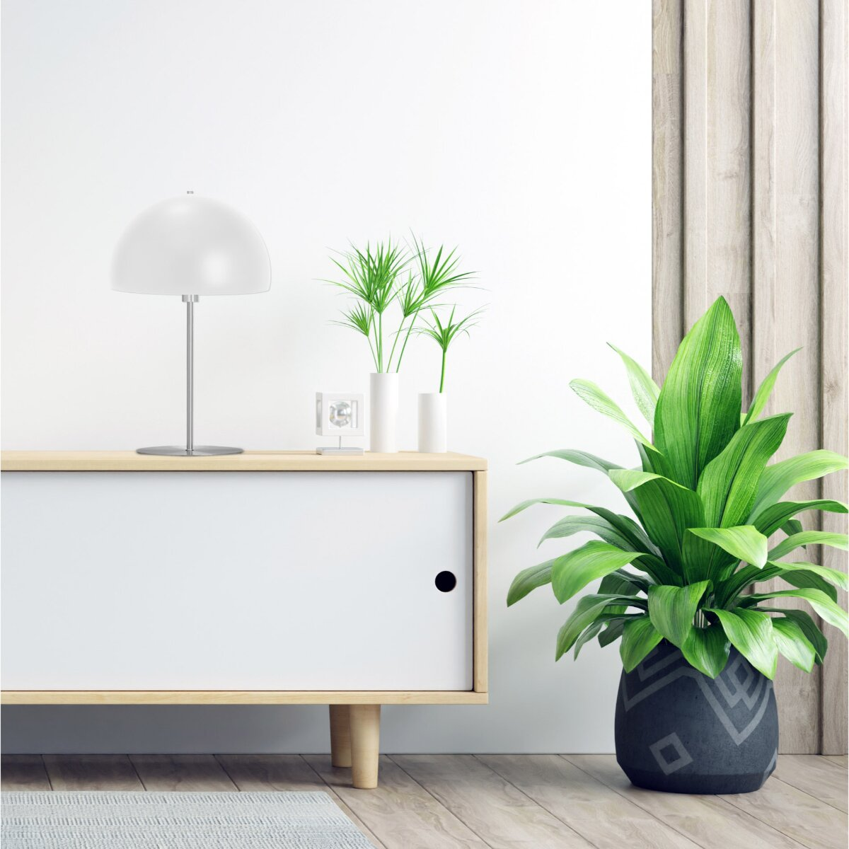 Eine weiße Lampe und grüne Pflanzen stehen auf einem hellen Holzschrank vor einer weißen Wand.