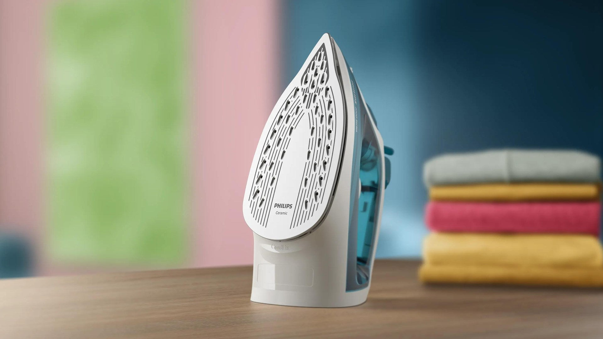 Un ferro da stiro Philips bianco e turchese su un tavolo di legno, con vestiti colorati sullo sfondo.