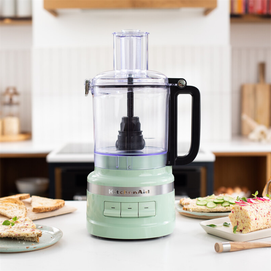 Fıstık yeşili bir KitchenAid mutfak robotu, aydınlık bir mutfakta beyaz tezgah üzerinde duruyor.
