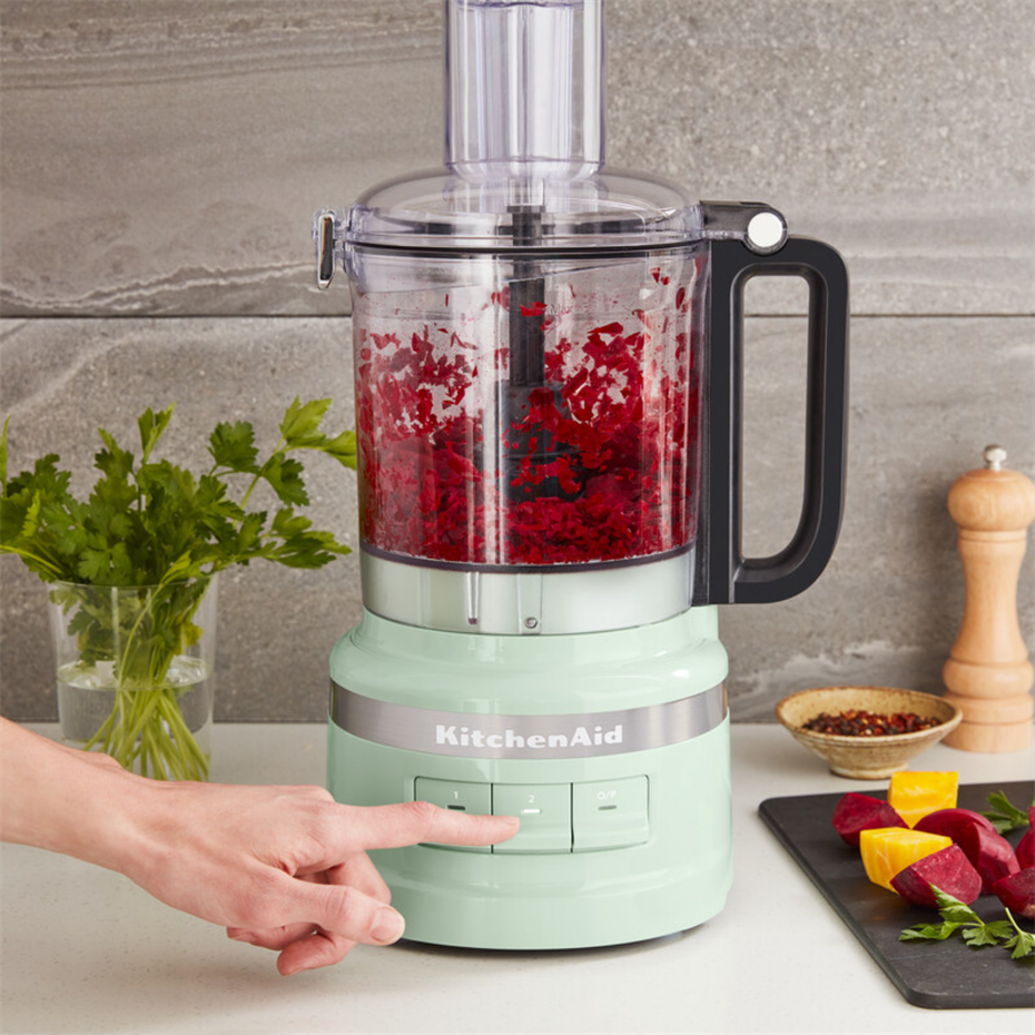 Bir el, doğranmış pancarla dolu nane yeşili bir KitchenAid mutfak robotunun düğmesine basıyor.