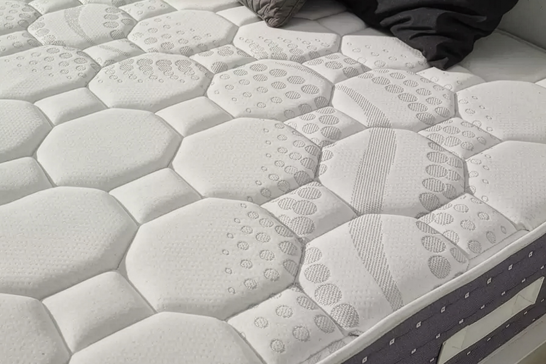 Egy fehér matrac geometriai steppelt mintával és sötét párnákkal a fejvégnél.
