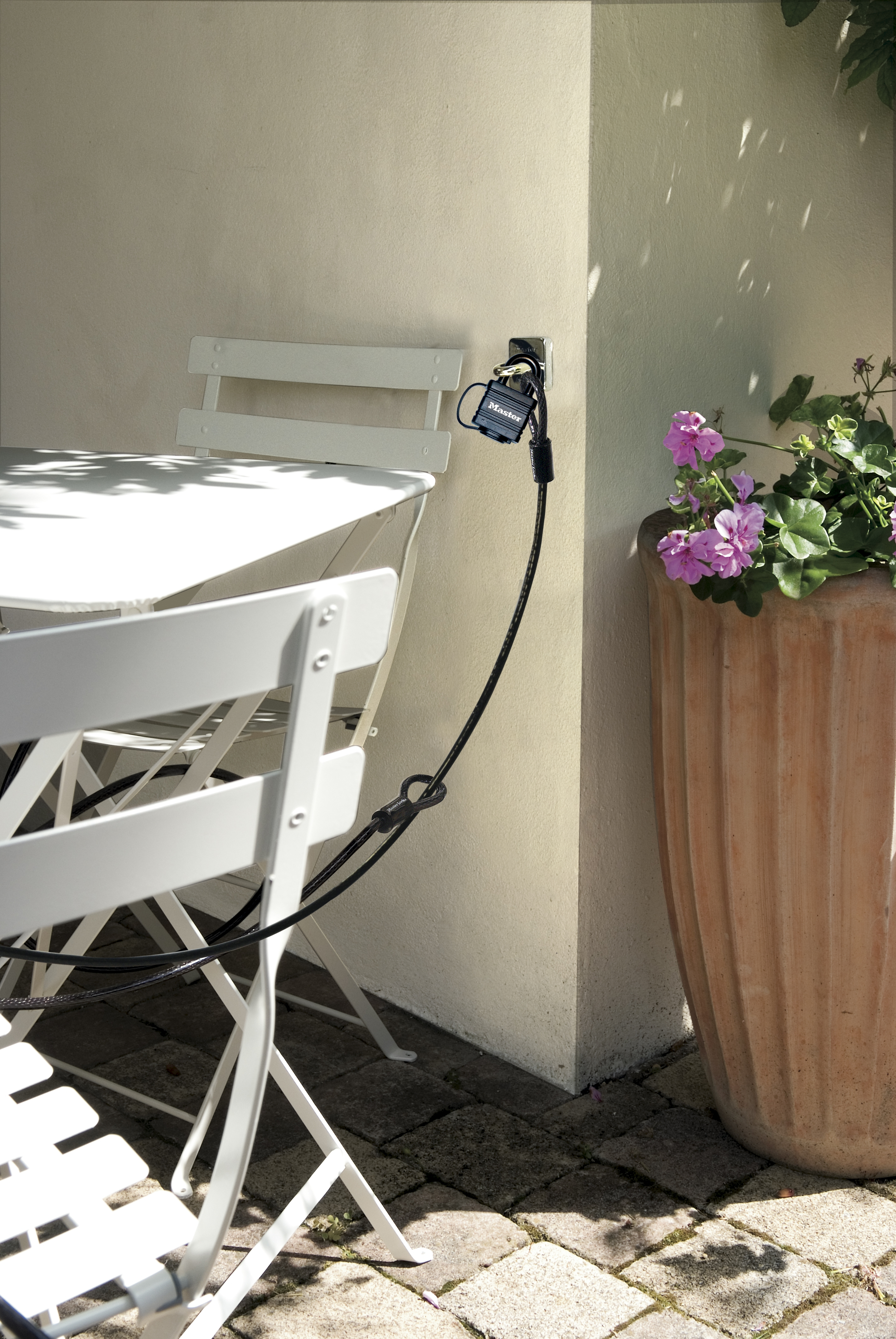 Ein weißer Tisch und Stühle stehen auf einer Terrasse, gesichert mit einem schwarzen Kabelschloss an der Wand.