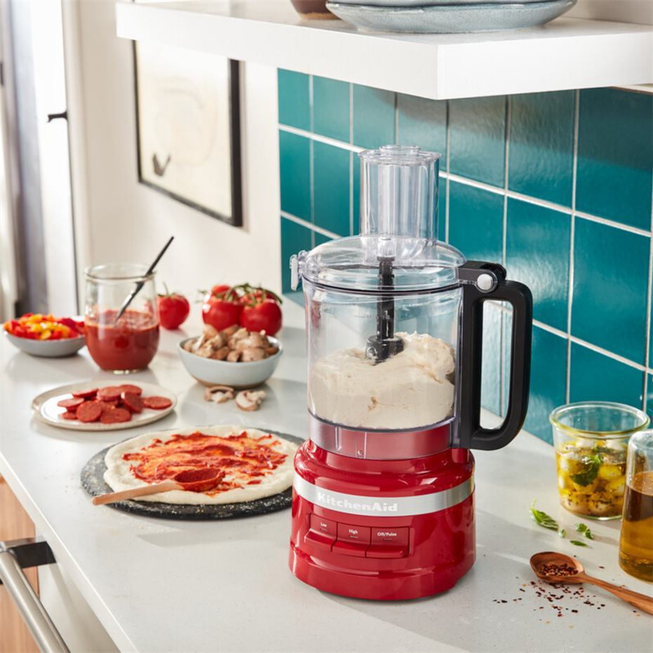 İçinde hamur olan kırmızı bir KitchenAid mutfak robotu tezgahta duruyor, yanında pizza malzemeleri var.