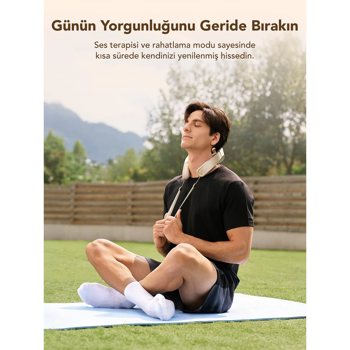 Bir adam yeşil bir manzarada boyun masaj aleti takarak açık havada meditasyon yapıyor.