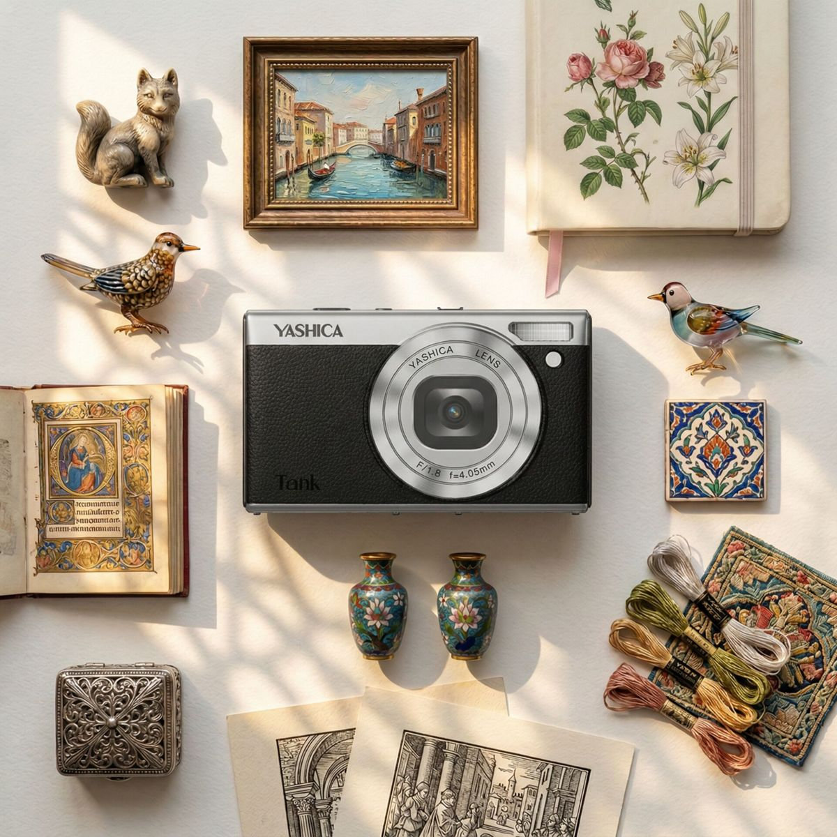 Eine Yashica-Kamera im Zentrum, umgeben von Kunst, Büchern, kleinen Figuren und Stickgarn auf hellem Hintergrund.
