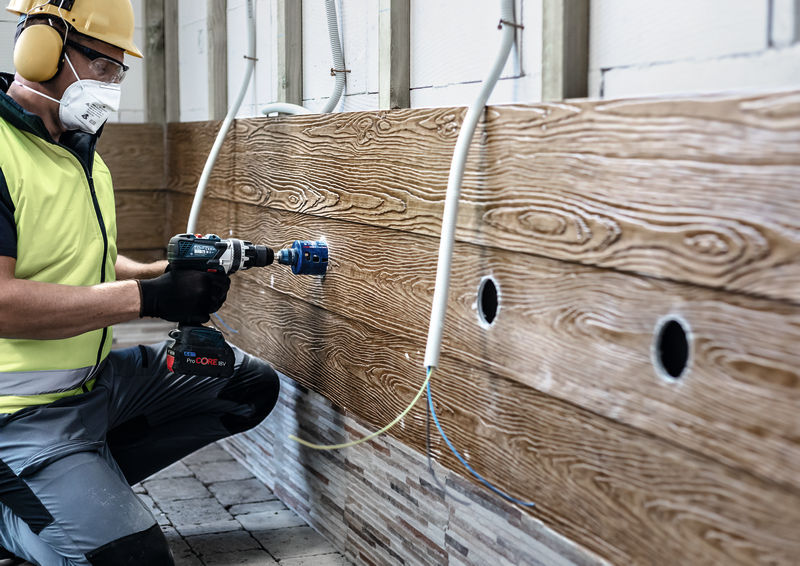 Ein Arbeiter mit Schutzkleidung bohrt Löcher in eine holzfarbene Wand.