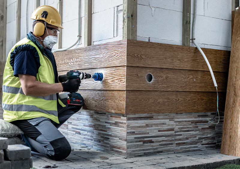 Ein Arbeiter mit Helm und Schutzbrille bohrt ein Loch in Holzverkleidung.