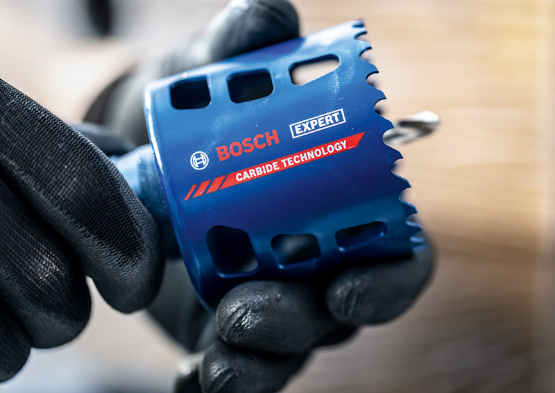 Bewehrte Hände halten eine blaue Bosch Expert Carbide Technology Lochsäge vor verschwommenem Hintergrund.