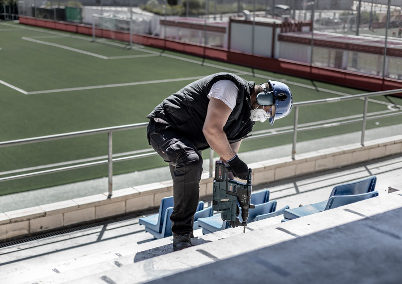 Güvenlik ekipmanlı bir işçi, yeşil saha manzaralı beton tribünlerde delik açıyor.
