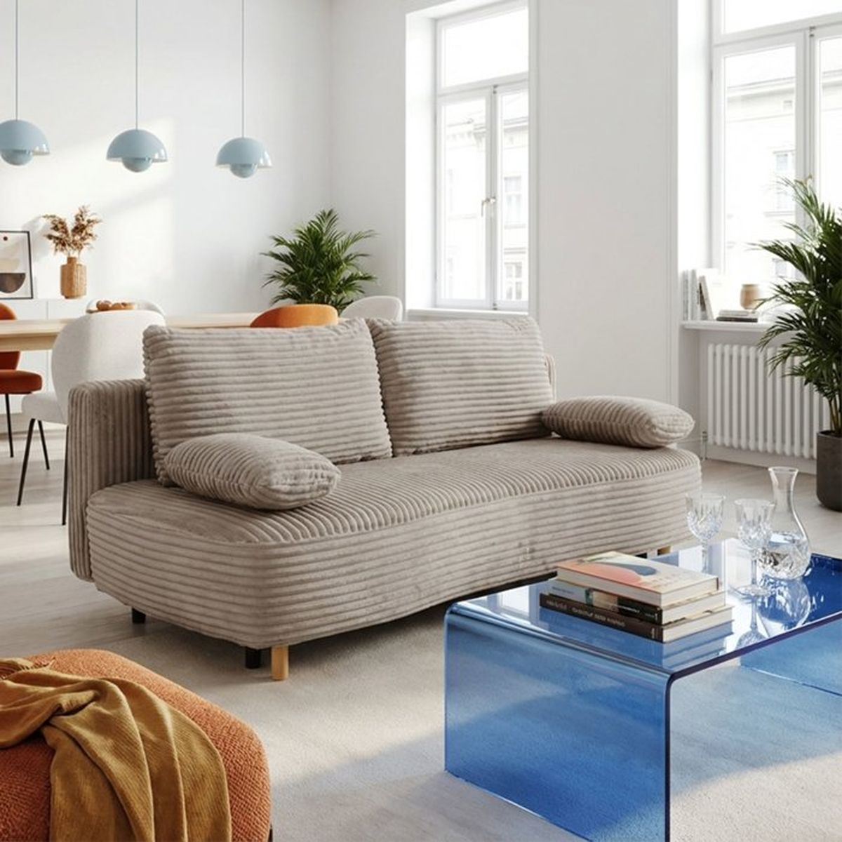 Ein beige Cordsofa in einem hellen Wohnzimmer mit blauem Glastisch, Pflanzen und Essbereich im Hintergrund.
