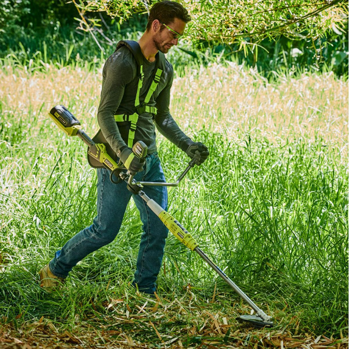 Ein Mann in Jeans und Gurt schneidet hohes Gras mit einem grünen Fadenträger.