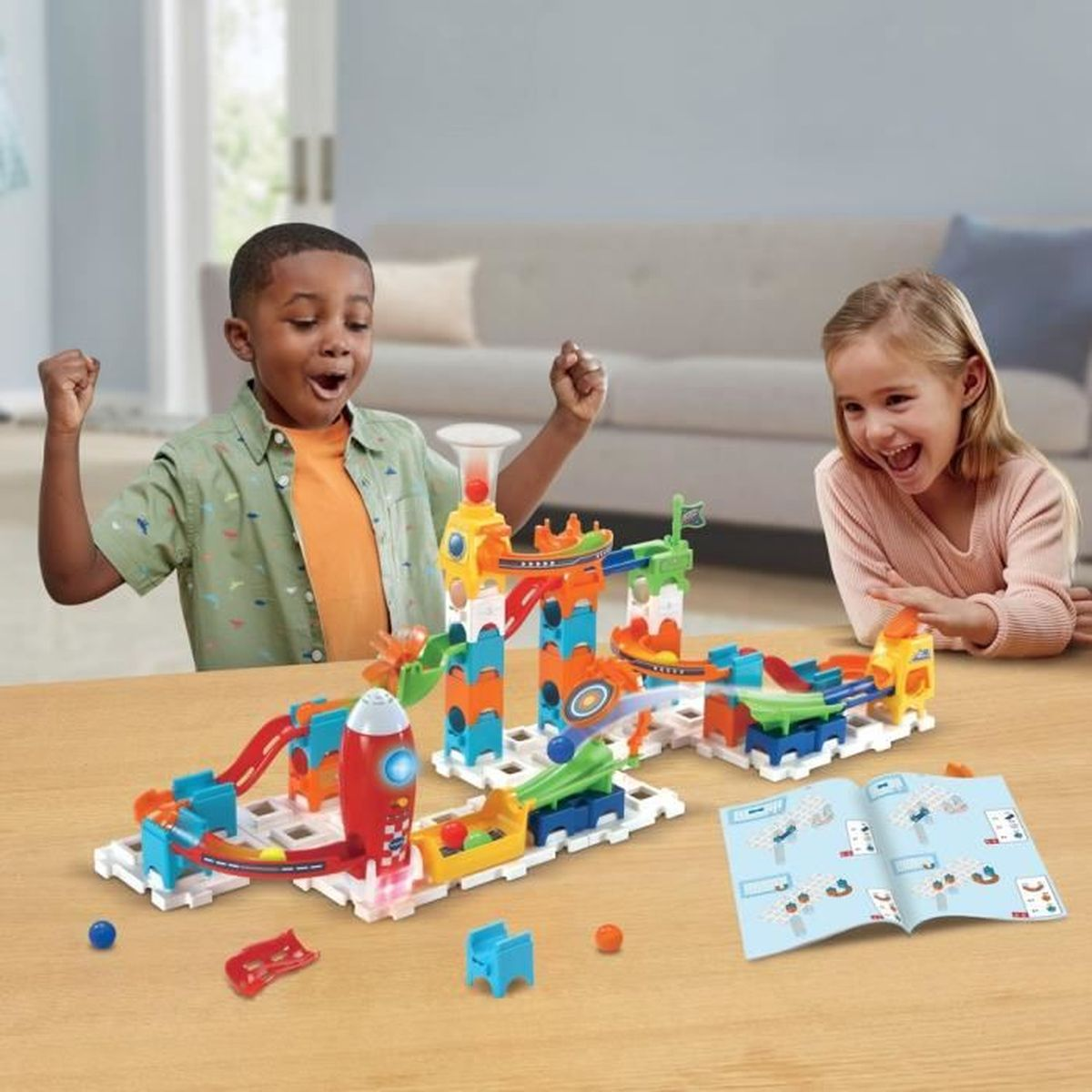 Zwei glückliche Kinder spielen am Tisch mit einer bunten Murmelbahn und einem Anleitungsbuch.