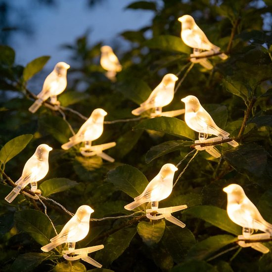Eine Lichterkette mit kleinen, weiß leuchtenden Vögeln hängt in grünen Büschen.