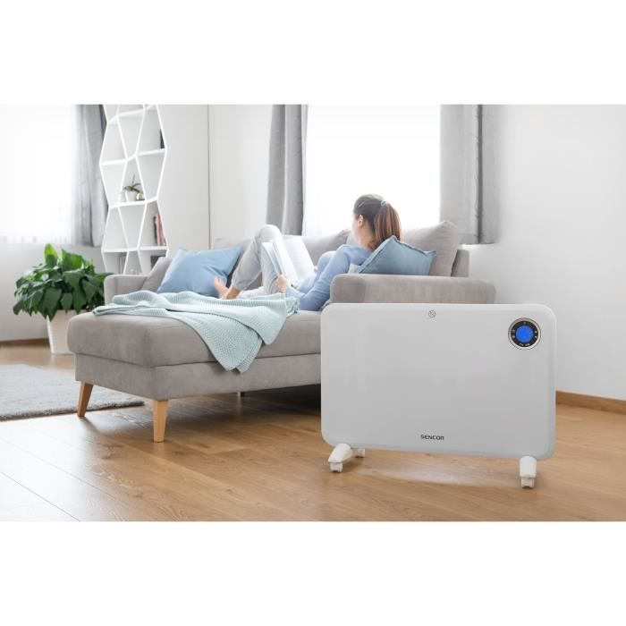 Ein weißer Elektroheizer steht auf einem Holzboden in einem Wohnzimmer, dahinter liest eine Frau auf dem Sofa ein Buch.