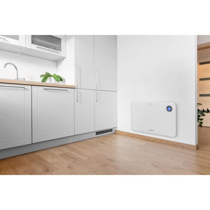 Ein weißer Wandkonvektor mit blauem Display hängt an einer weißen Wand neben einer modernen weißen Küche.