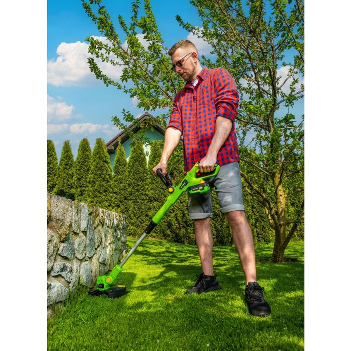 Ein Mann in rot-kariertem Hemd trimmt Gras mit einem grünen Rasentrimmer im Garten.