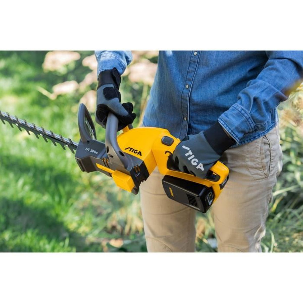 Person mit Handschuhen hält eine gelbe Stiga-Heckenschere in einem Garten.