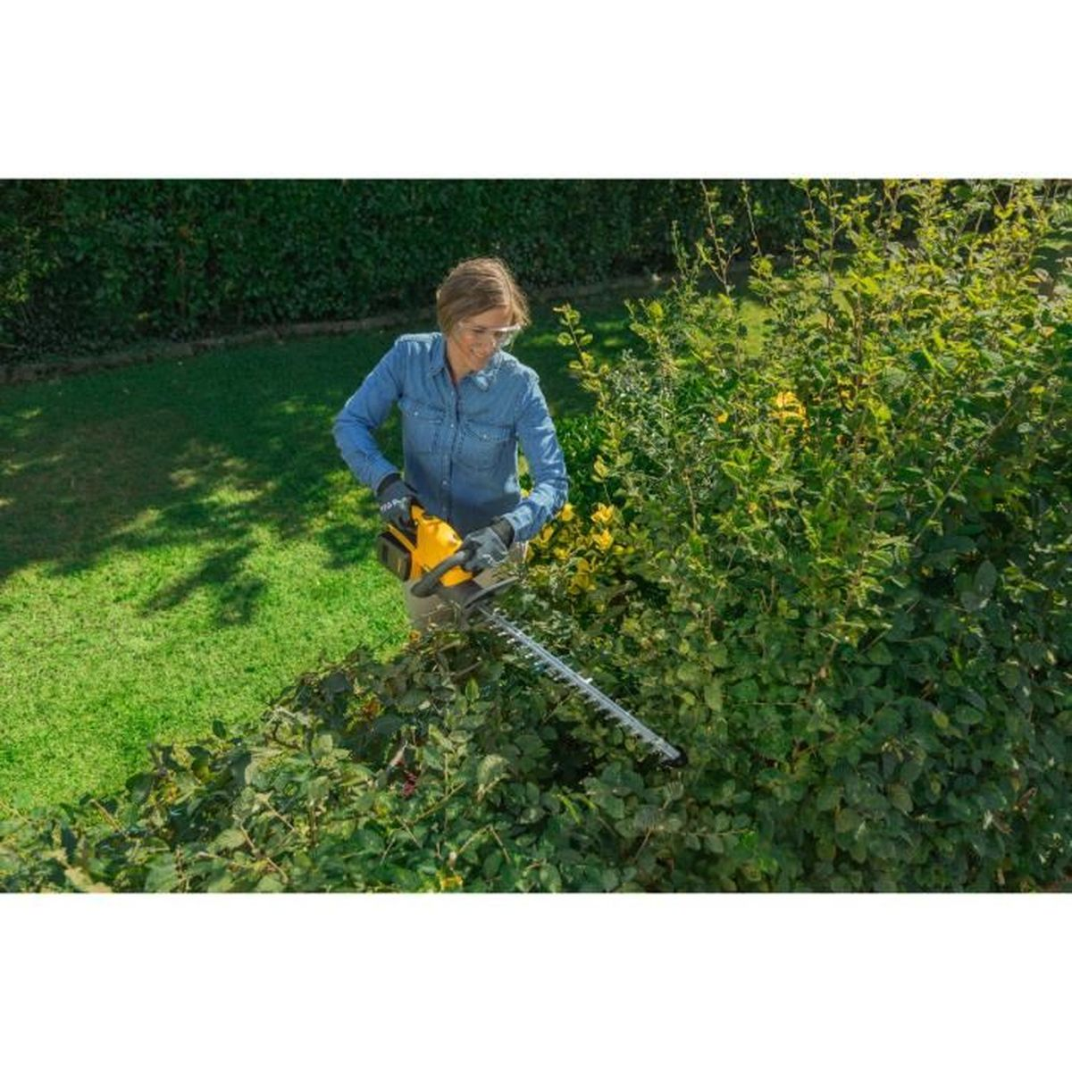 Frau in Jeanshemd schneidet grüne Hecke mit einer elektrischen Heckenschere im Garten.