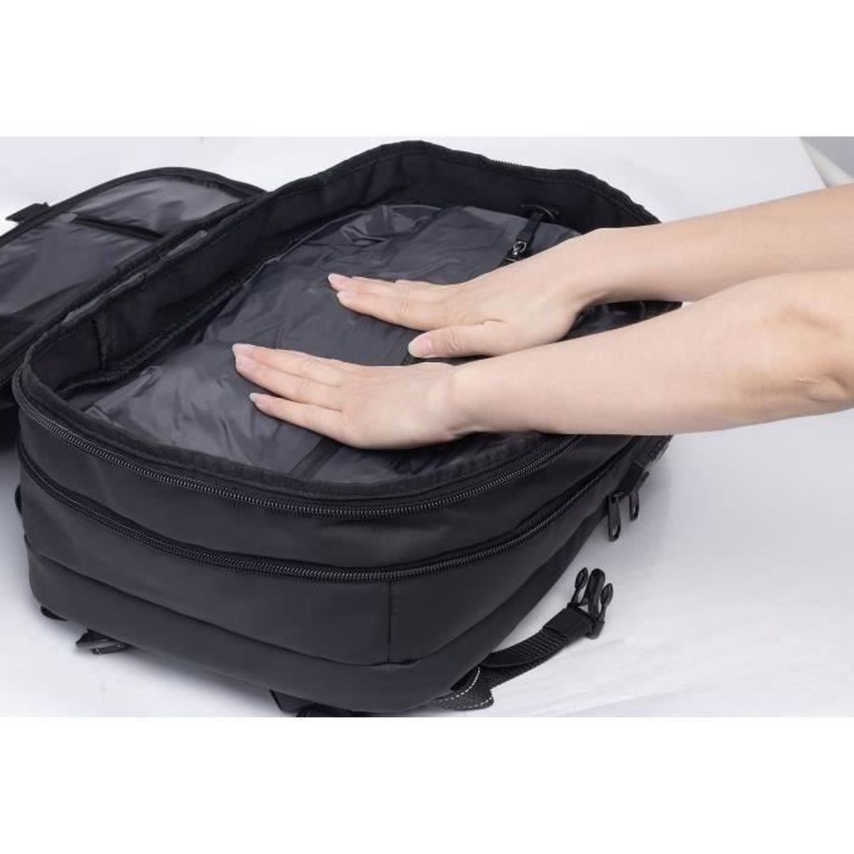 Hände drücken Kleidung in einem offenen schwarzen Rucksack auf weißem Grund.