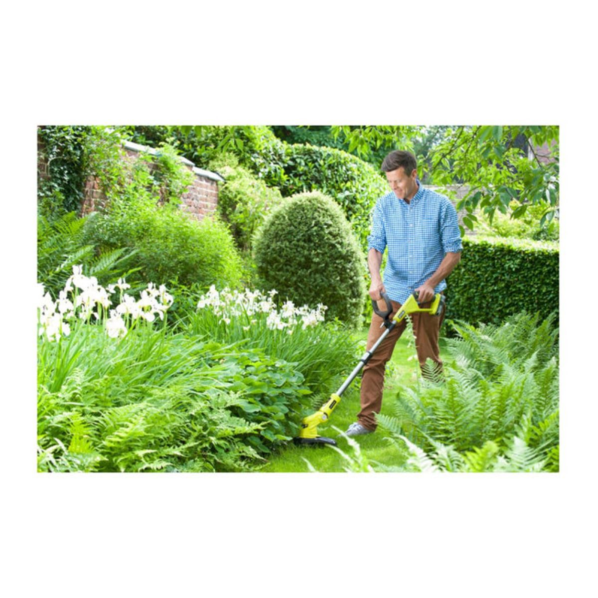 Ein Mann in blauem Hemd und brauner Hose benutzt einen Rasentrimmer in einem üppigen Garten.