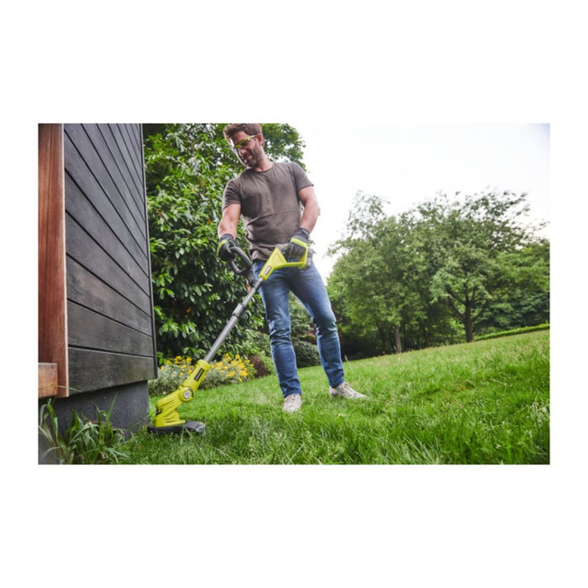 Ein Mann in Jeans und grauem Shirt benutzt einen grünen Rasentrimmer im Garten.