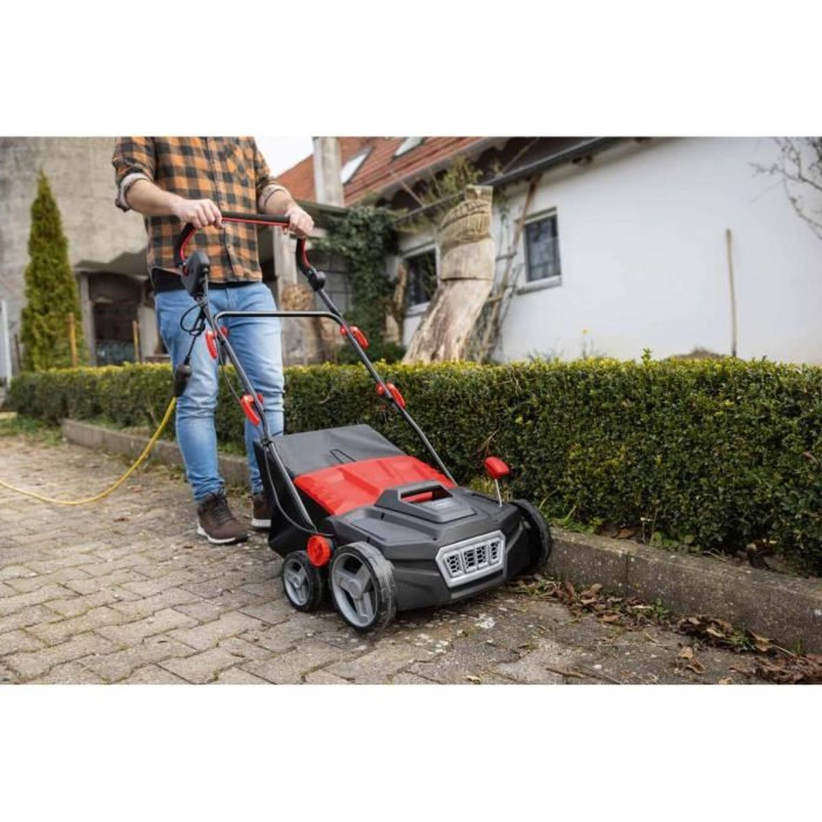 Ein Mann im karierten Hemd bedient einen roten und schwarzen Rasenlüfter auf einem gepflasterten Weg neben einer Hecke.
