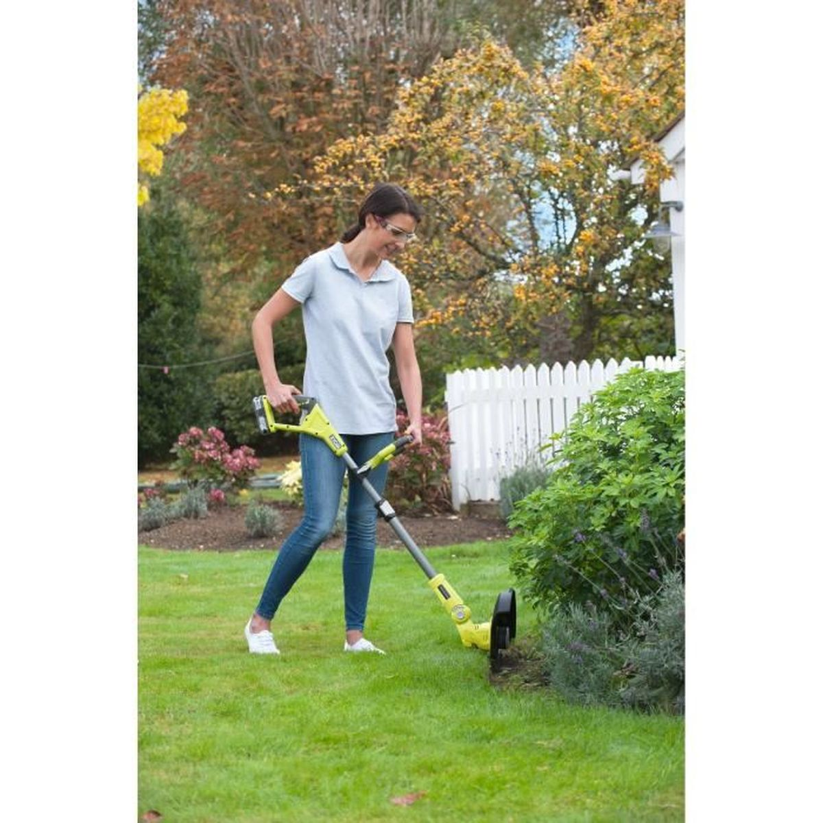 Eine Frau in einem hellen Hemd und Jeans trimmt mit einem Rasentrimmer die Rasenkante in einem Garten.
