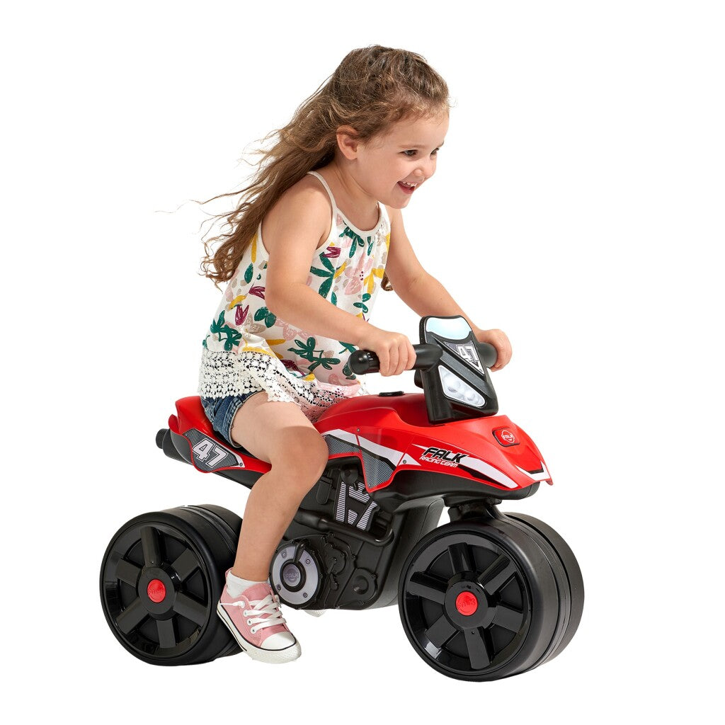 Ein kleines Mädchen mit braunen Haaren fährt auf einem roten Spielzeug-Motorrad mit breiten schwarzen Rädern.