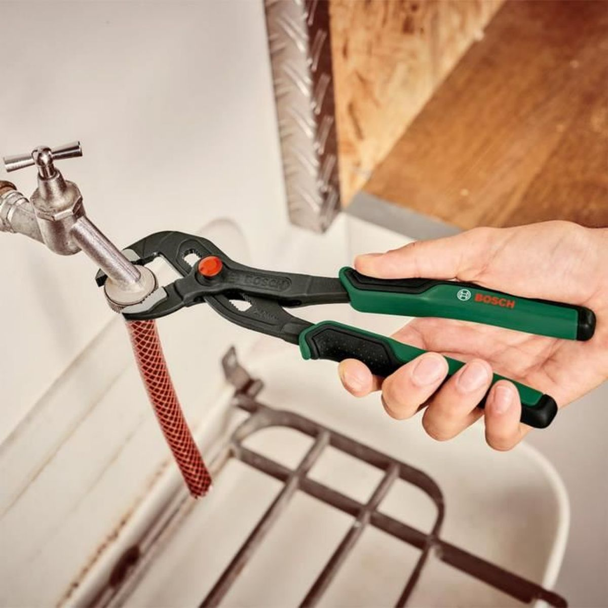Eine Hand hält eine Bosch-Wasserpumpenzange, um eine Armatur mit angeschlossenem rotem Schlauch zu greifen.