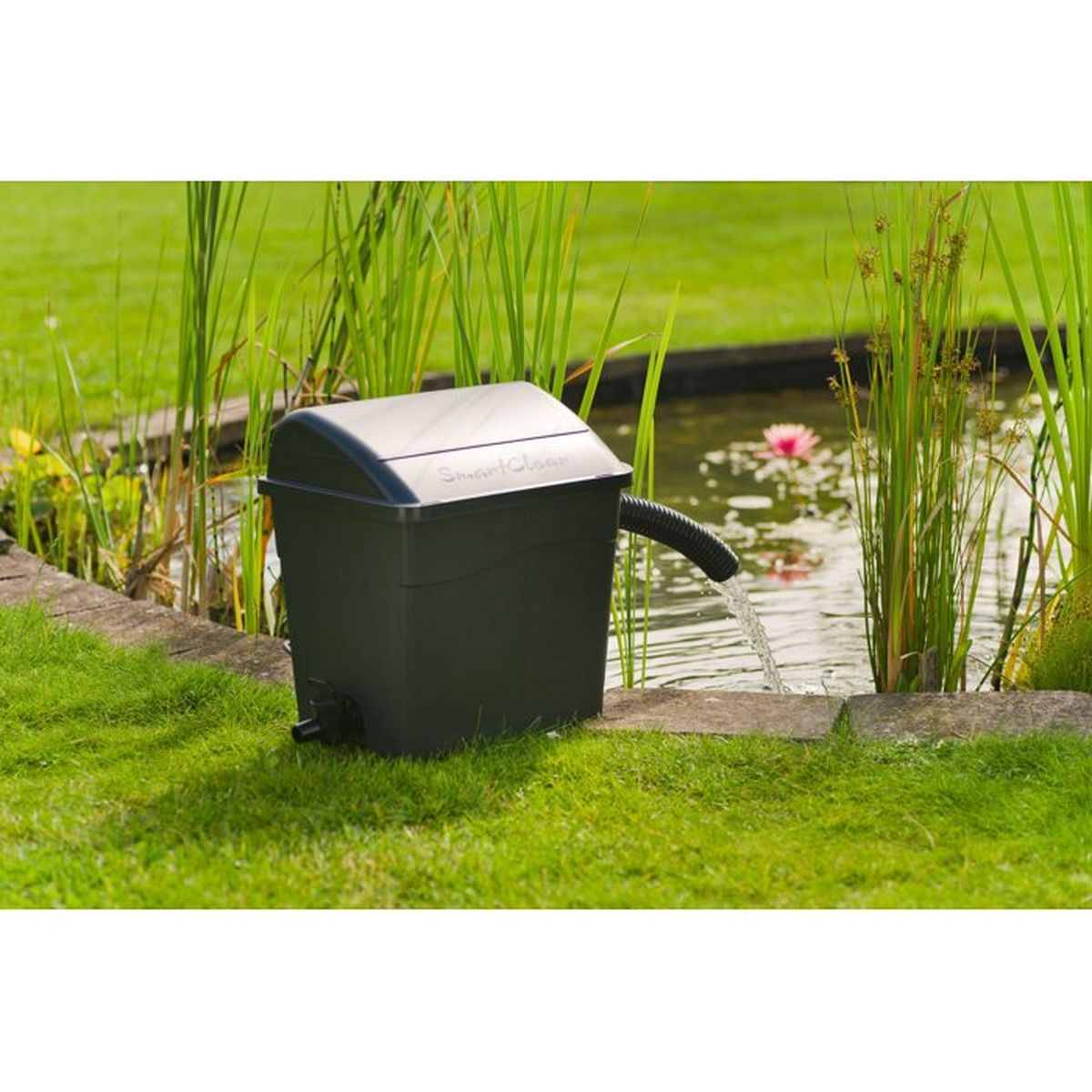 Ein grauer Teichfilter SmartClear steht am grünen Ufer eines Gartenteichs und leitet Wasser zurück.