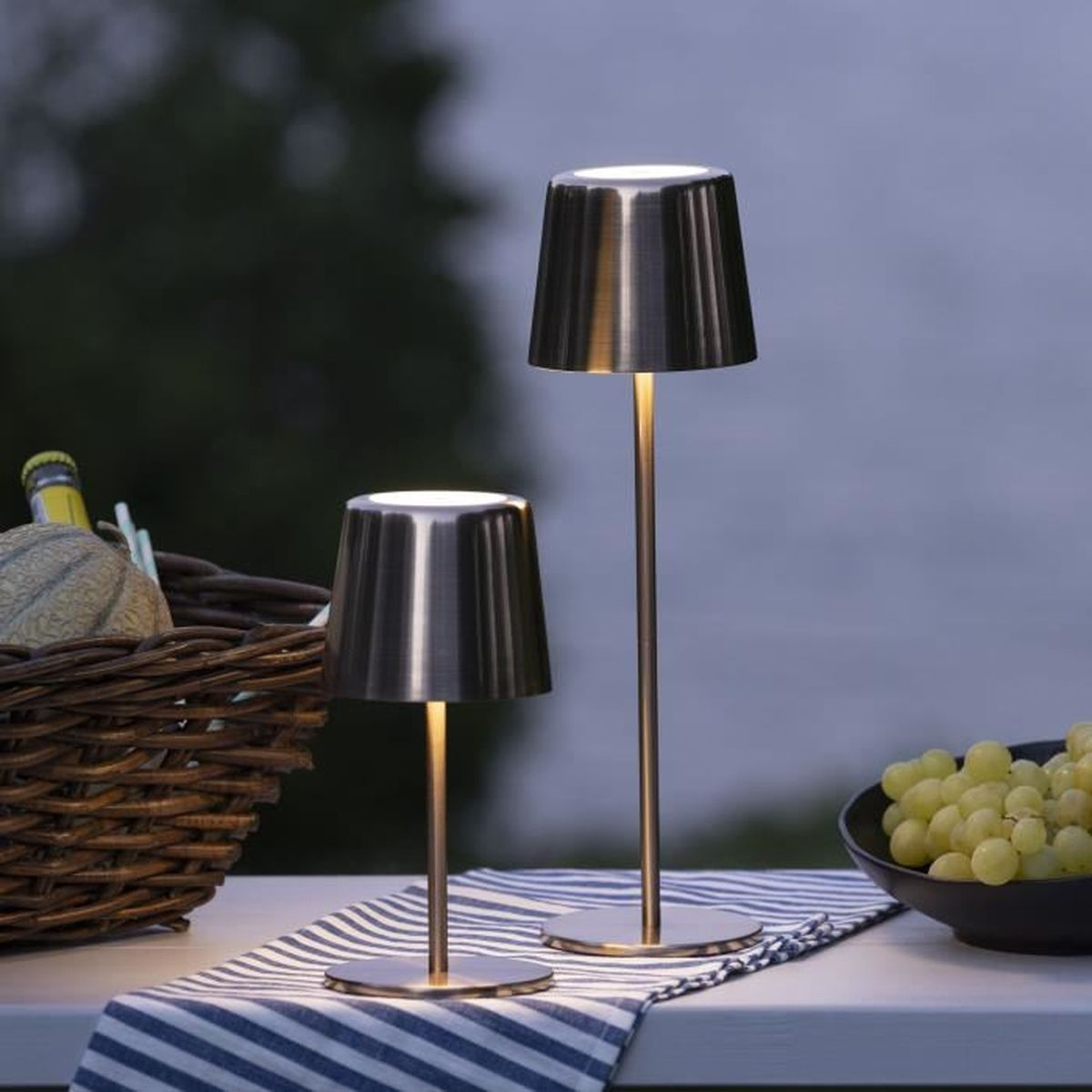 Zwei Metalltischlampen auf einem gestreiften Läufer, mit einem Weidenkorb und grünen Trauben auf einem Tisch im Freien bei Dämmerung.