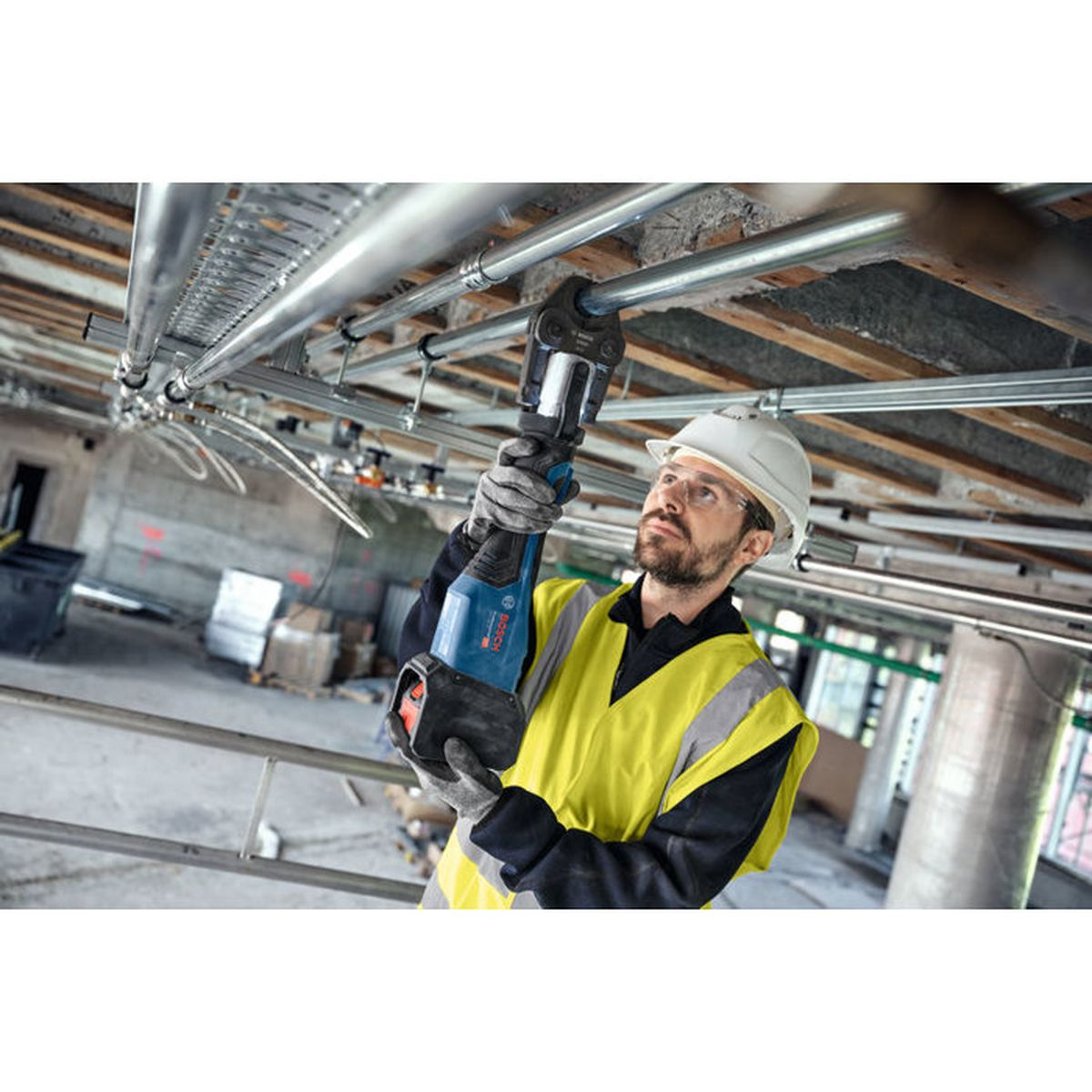 Ein Arbeiter in gelber Weste und Schutzhelm presst Rohre an einer Decke mit einem Bosch-Werkzeug.