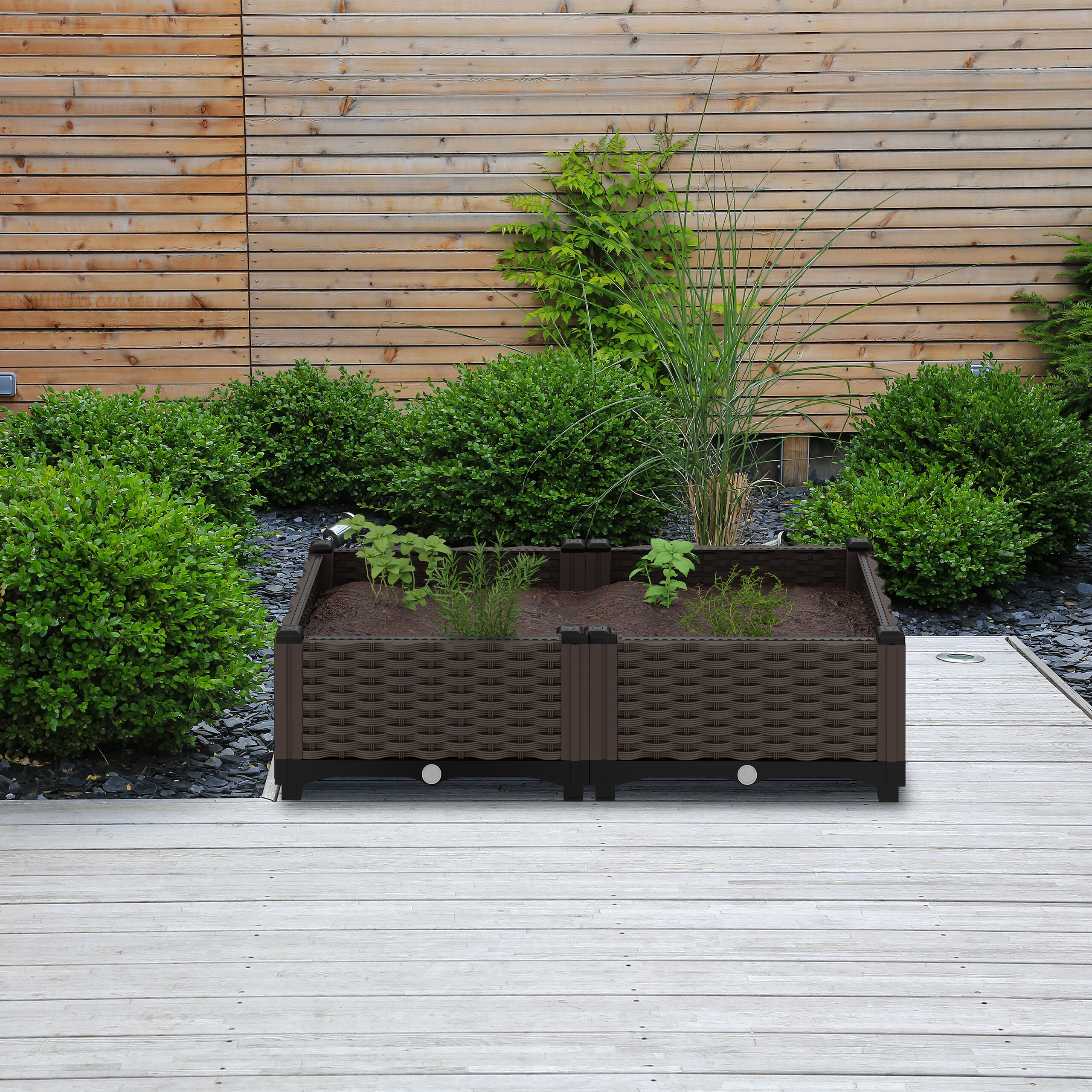 Ein braunes Rattan-Hochbeet steht auf einer hellen Holzterrasse vor einer Holzwand und grünen Büschen.
