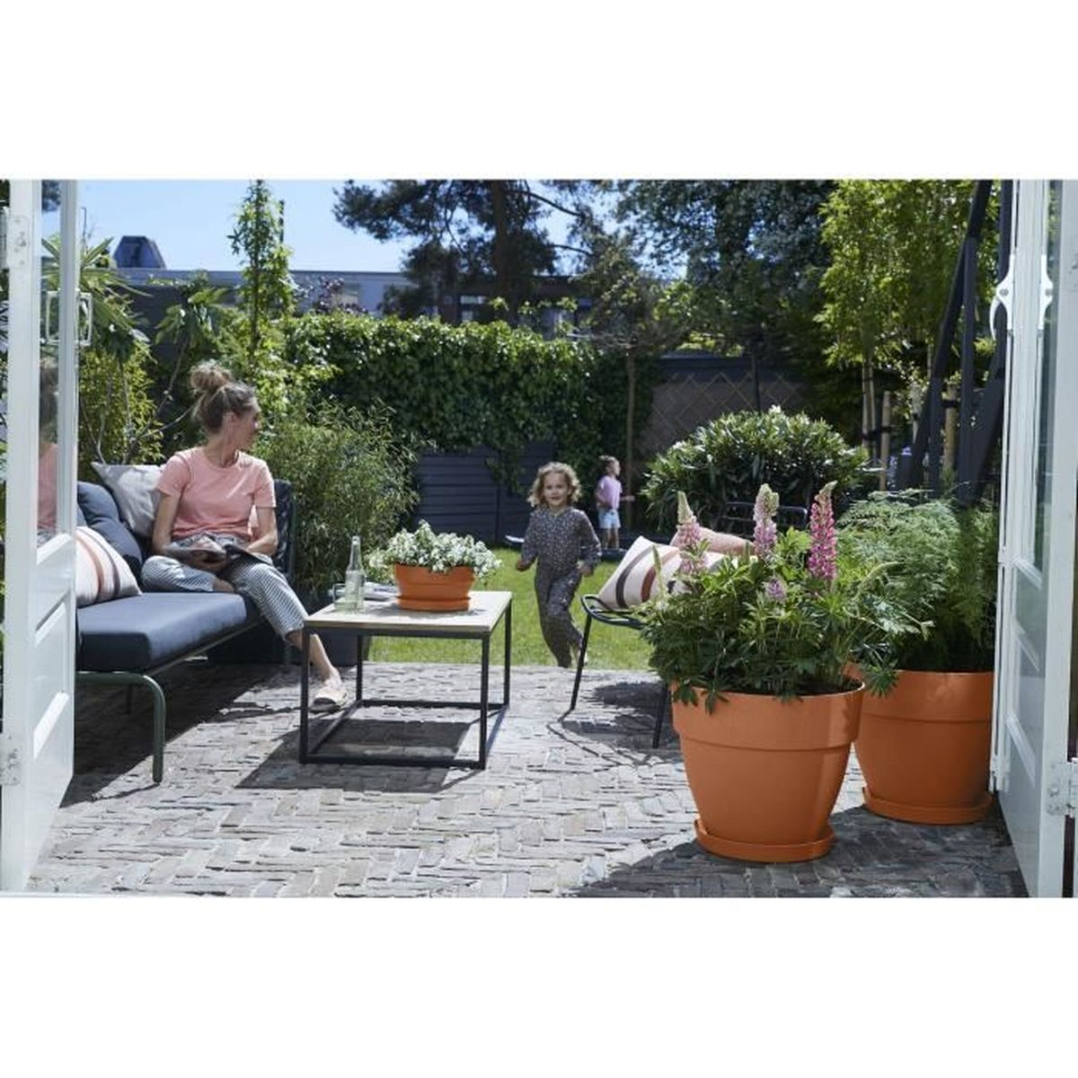 Eine Frau sitzt auf einer Gartenbank auf einer Steinterrasse, während ein Kind im Garten im Hintergrund spielt.