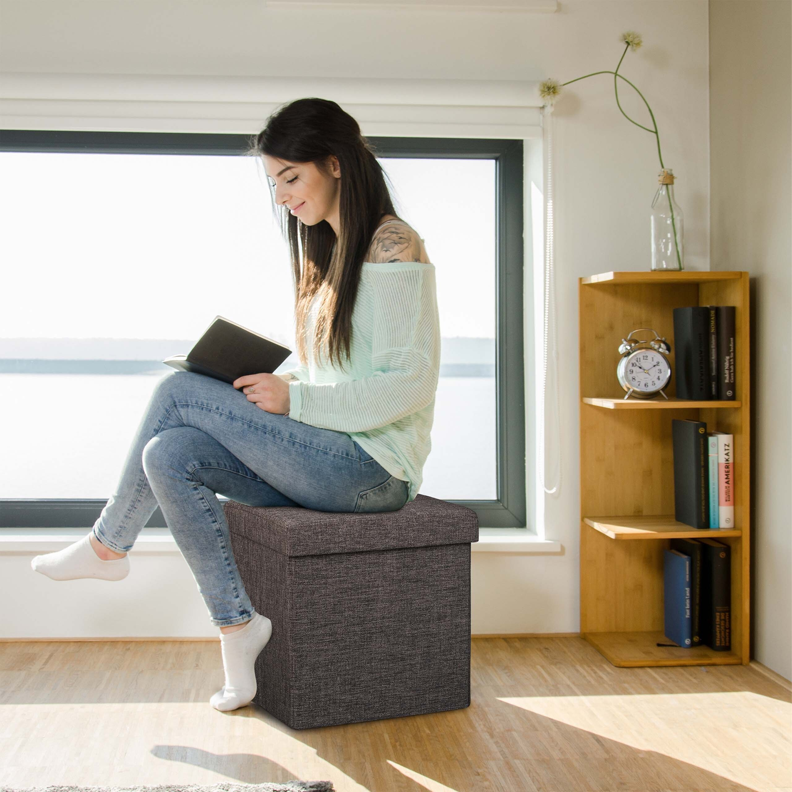 Eine Frau mit langem dunklem Haar sitzt auf einem grauen Hocker vor einem Fenster und liest ein Buch.