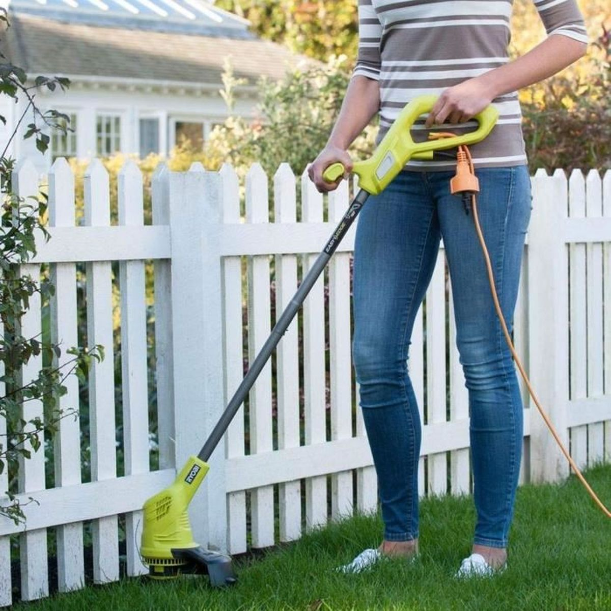 Eine Frau im Garten nutzt einen gelben Rasentrimmer entlang eines weißen Zauns.
