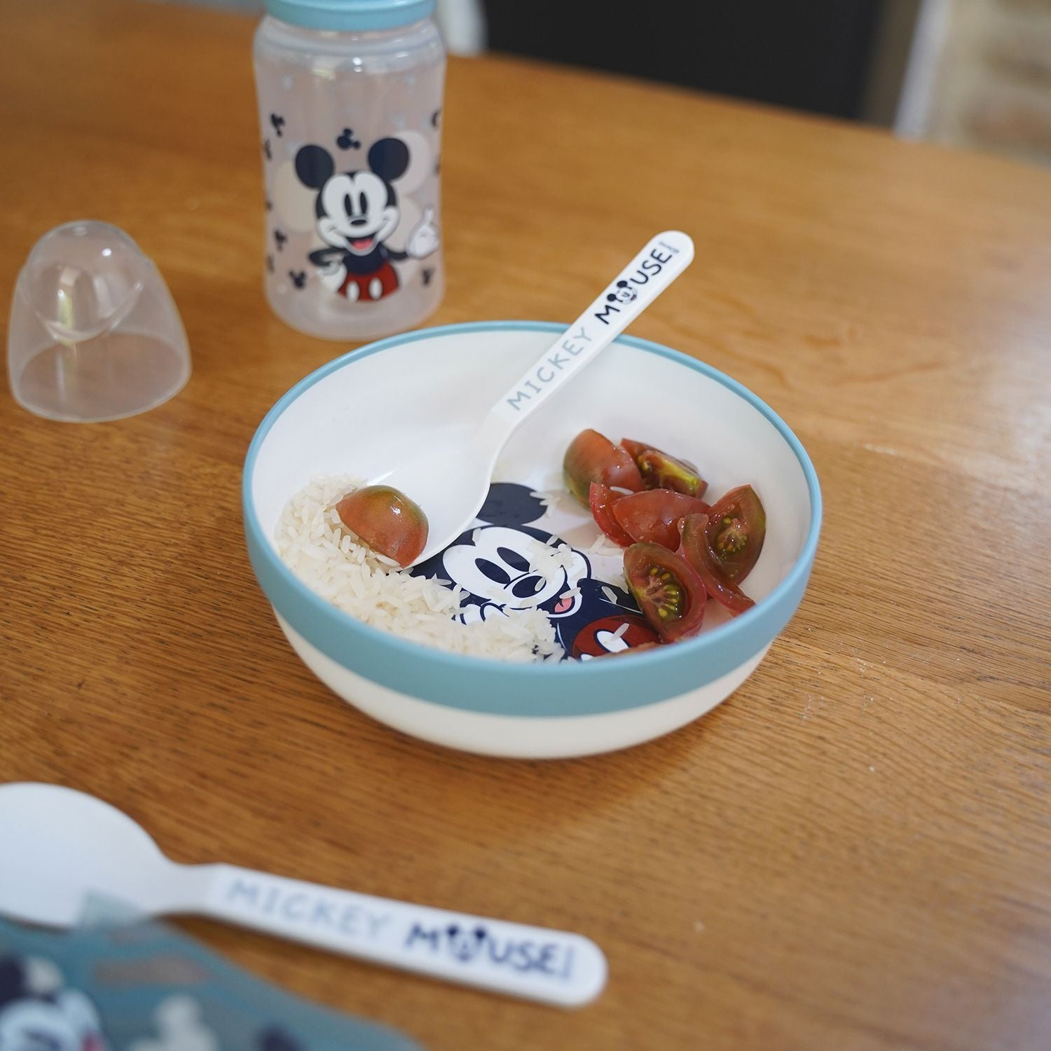 Mickey-Maus-Schüssel mit Reis und Tomaten auf Holztisch, daneben Löffel und Flasche.