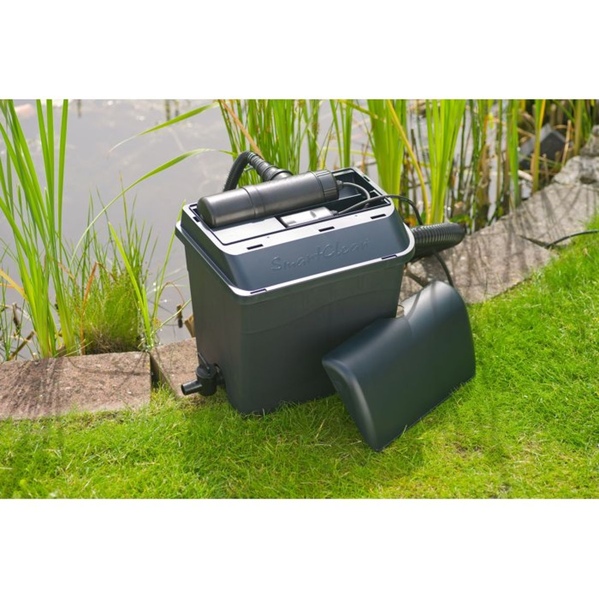 Ein grauer Gartenteichfilter auf grünem Rasen neben einem Teich mit Schilf und einem abgenommenen Deckel.