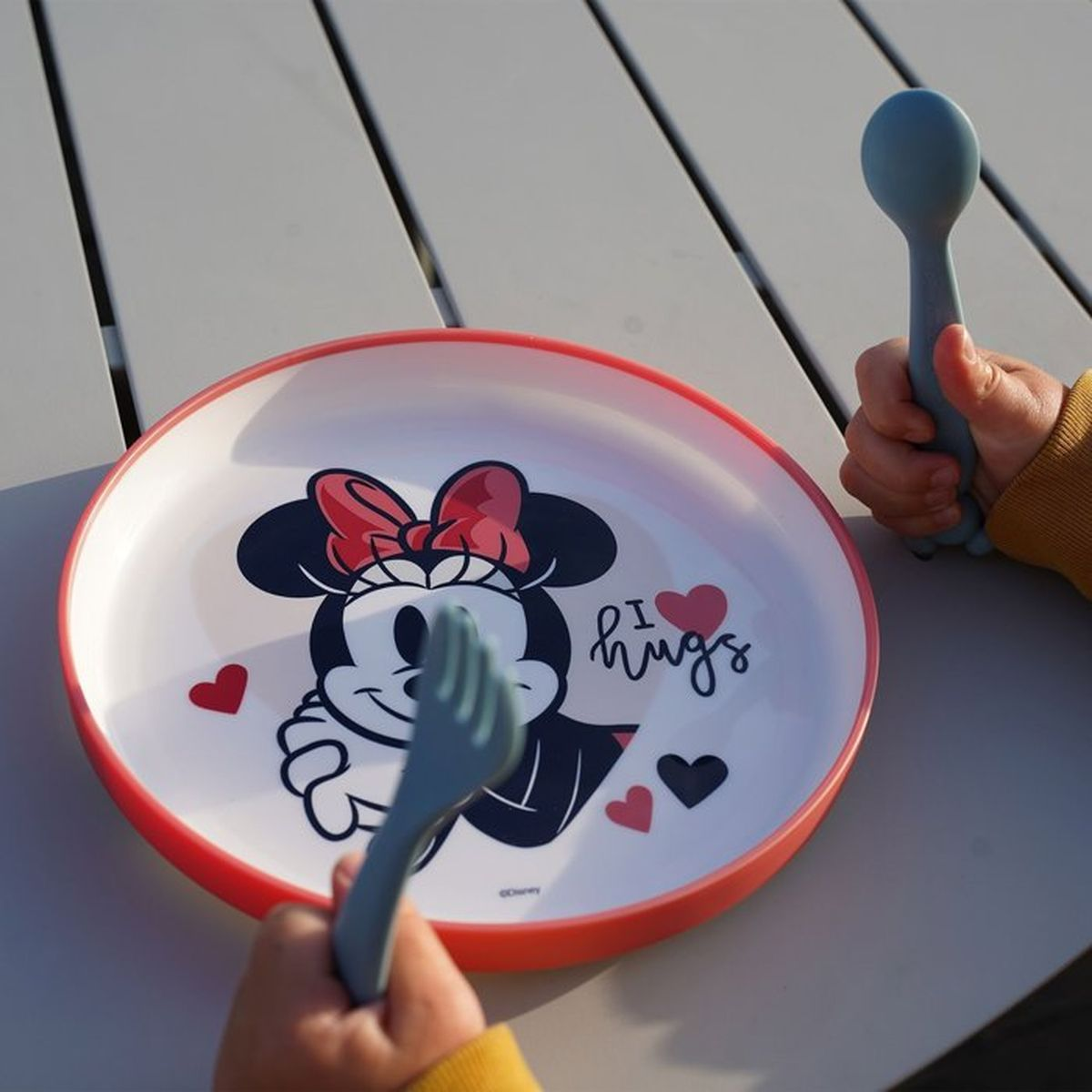 Ein Kind hält eine blaue Gabel und einen Löffel vor einem Minnie-Maus-Teller.
