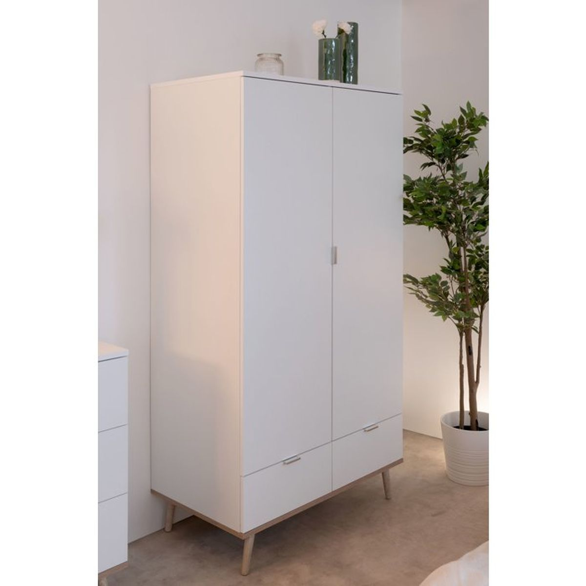 Ein weißer Kleiderschrank mit Holzakzenten und Beinen steht vor einer weißen Wand, rechts daneben eine Topfpflanze.