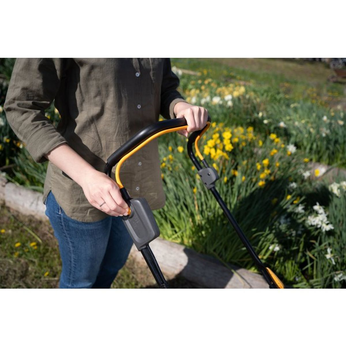 Eine Frau hält die Griffe eines Gartenwerkzeugs vor einem Garten mit gelben Blumen.