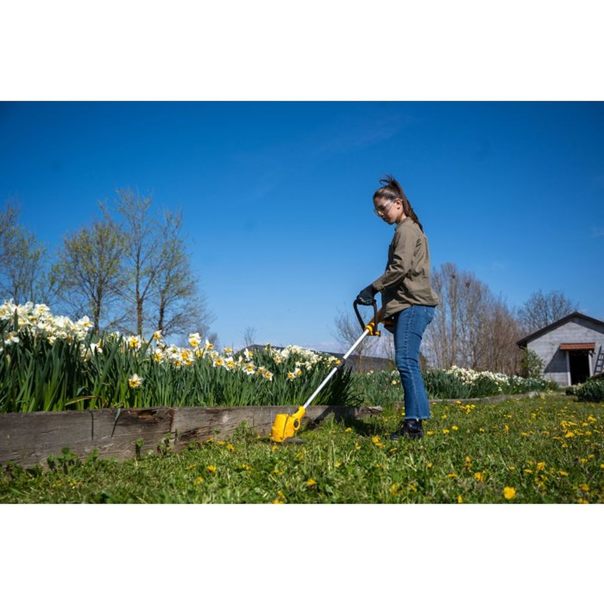 Eine Frau nutzt einen Rasentrimmer in einem Garten mit Blumenbett und blauem Himmel.